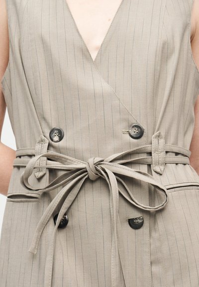 Μπεζ γιλέκο χωρίς μανίκια με λεπτή ρίγα, διπλό κουμπί και δεμ ένη ζώνη από ύφασμα στη μέση, εμφανιζόμενο στον κορμό ενός ατόμου.