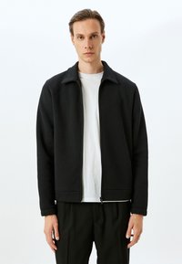 Chaqueta negra con cremallera y cuello alzado, hecha de tejido texturizado. El diseño presenta un corte recto y puños acanalados, llevada sobre una camisa blanca.