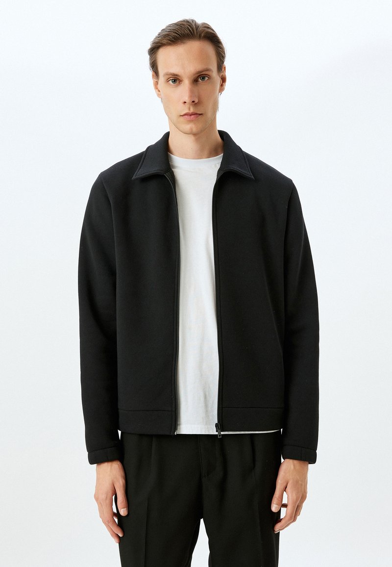 Chaqueta negra con cremallera y cuello alzado, hecha de tejido texturizado. El diseño presenta un corte recto y puños acanalados, llevada sobre una camisa blanca.