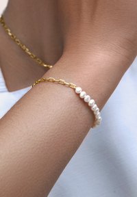 Goldene Kettenarmband mit ovalen Gliedern, kombiniert mit einer kleinen Reihe weißer irideszenter Perlen. Sanfte Textur auf der Haut.