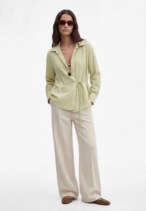 Kobieta ubrana w jasnooliwkową bluzkę zapinaną na zakładkę, beżowe spodnie o szerokich nogawkach, brązowe buty, warstwowe naszyjniki i brązowe okulary przeciwsłoneczne, stoi z rękami w kieszeniach.