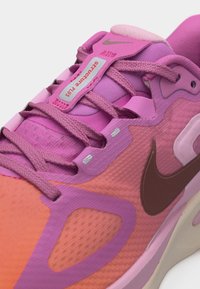 Κοντινή λήψη ενός ροζ και πορτοκαλί παπουτσιού τρεξίματος Nike με ροζ κορδόνια και ετικέτα που γράφει «Structure Plus» στη γλώσσα.