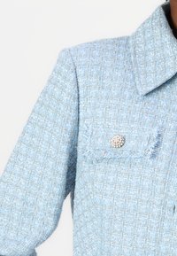 Vaaleansininen tweed-takki, jossa on kaulus ja yksi rintatasku, koristeltuna pyöreällä, koristellulla hopeanvärisellä napilla. Rakenteellinen kangas.