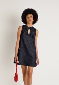 Denim šatová sukňa bez rukávov s detailom mašle, výstrihom v tvare kľúča a rozptýlenými bielymi ozdobami. Model drží malú červenú kabelku.
