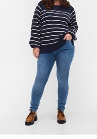 Námořnický a krémový pruhovaný svetr s volnými rukávy, kombinovaný s těsnými modrými džíny a hnědými šněrovacími botami. Texturovaný pletený materiál a uvolněný střih.