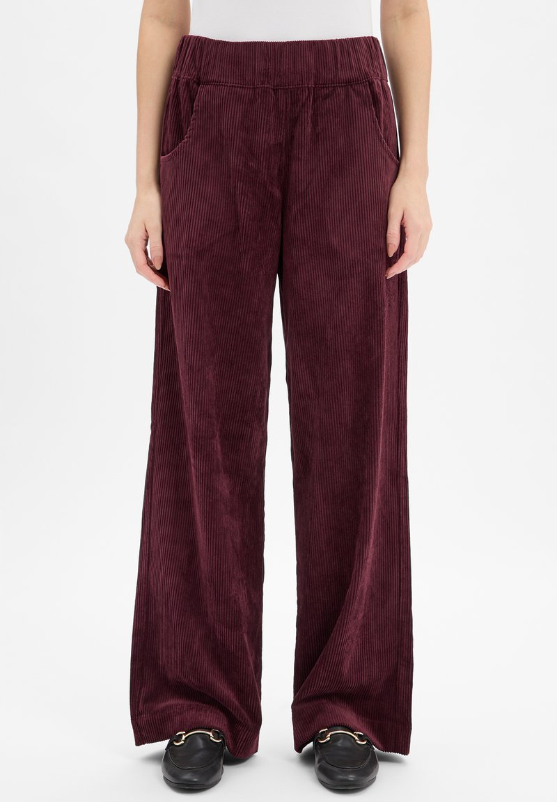 Pantalons en velours côtelé bordeaux à jambes larges avec une taille élastique, des poches latérales et une texture à côtes verticales, associés à des mocassins noirs.
