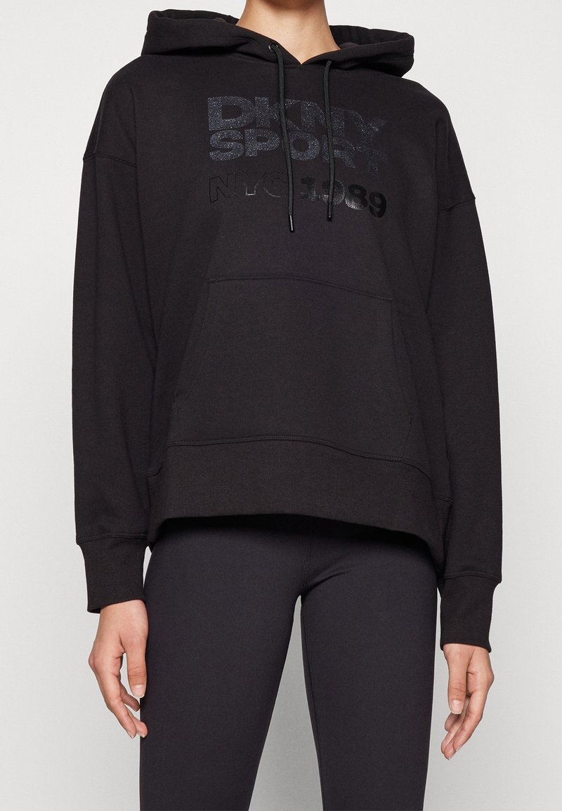 Sweat à capuche noir oversize avec le texte "DKNY SPORT NYC 1989", associé à un legging noir ajusté sur une personne debout.