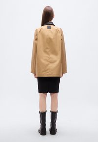 Μπεζ εξωτερικό μπουφάν με μαύρο γιακά, που διαθέτει ορθογώνια ετικέτα στην πλάτη. Φορώντας το πάνω από μια μαύρη φούστα, σε συνδυασμό με μαύρες μπότες μέχρι το γόνατο.