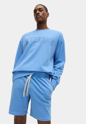 Młody mężczyzna w jasnoniebieskiej bluzie z napisem „WEEKEND” oraz dopasowanych szortach z białym sznurkiem, stojący na jednolitym tle.