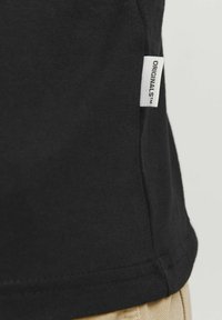 Zwarte katoenen t-shirt met een rechte snit, met een witte stoflabel met de tekst "ORIGINALS™" op de onderste zijnaad.