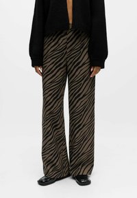 Pantalons larges avec des rayures zèbre marron et noir, associés à des chaussures noires brillantes à bout carré et un pull noir avec des manches visibles.