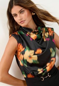 Top de satén negro con patrón floral en naranja y verde, cuello drapeado, diseño sin mangas, combinado con pantalones de talle alto con herrajes plateados.