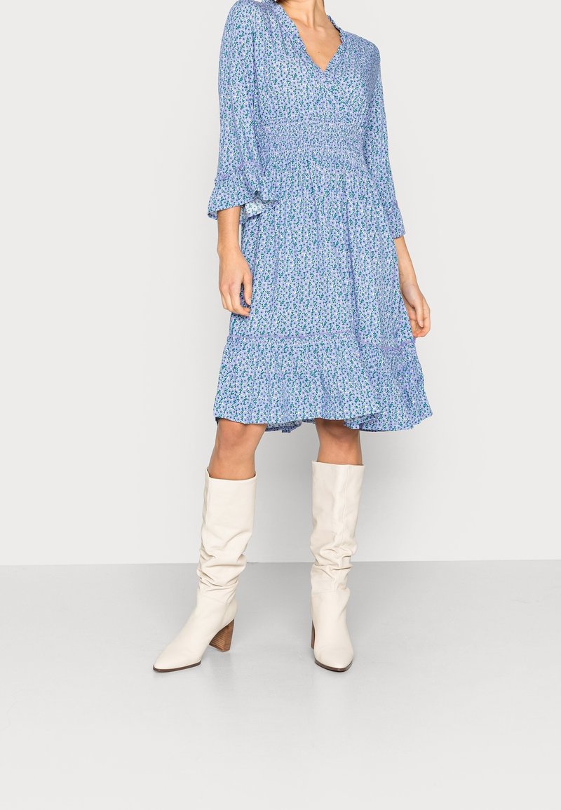 Modrá květinová šaty s výstřihem do V, tříčtvrteční volánkové rukávy a sukni se střídavými vrsty. Nosit se bílými koženými holínkami po kolena a s vysokým podpatkem.