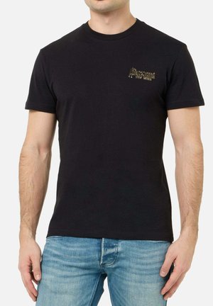 Man wearing a plain black short-sleeve t-shirt with a small gold "Boxeur Des Rues" logo on the left chest, paired with blue jeans.
