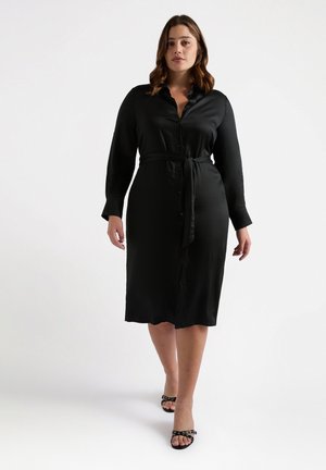 Robe chemise noire à manches longues avec un devant à boutons, une ceinture assortie à la taille, et un tissu lisse et brillant. Portée avec des talons ouverts.