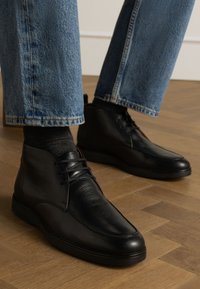 Bottes mi-mollet en cuir noir avec une texture lisse, dotées de lacets et d'un bout arrondi. Portées avec un pantalon en denim bleu clair. Sol en bois en dessous.