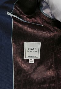 Wnętrze granatowego garniturowego żakietu z geometrycznym wzorem w burgundzie, z białą metką z informacjami o marce i rozmiarze. Akcentowane jasnoniebieskimi wykończeniami.