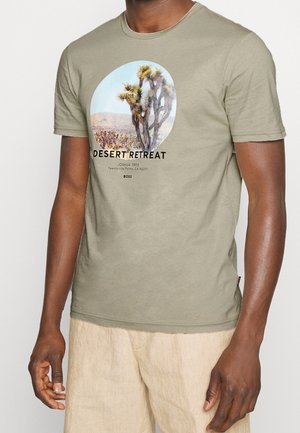 Mężczyzna noszący jasnorozmarynowy t-shirt z grafiką pustyni Joshua Tree oraz napisem „Desert Retreat” z przodu, w zestawie z beżowymi spodniami.