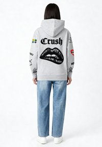 Grijze hoodie met een grafisch ontwerp van lippen en het woord "Crush" in dikke letters op de achterkant, met kleurrijke patches op de mouwen.