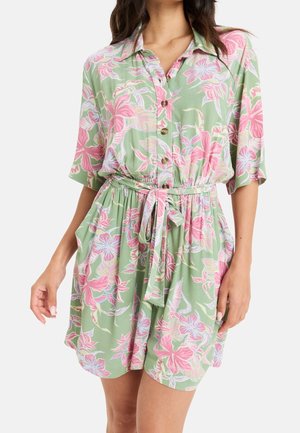 Vrouw draagt een groene jurk met korte mouwen en knopen, met roze en paarse bloemenprint en een stoffen riem gestrikt in de taille.