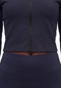 Prim-plan cu o persoană care poartă un top închis la culoare, plisat, cu fermoar și colanți înalți asortați, evidențiind un mic spațiu la mijlocul abdomenului.