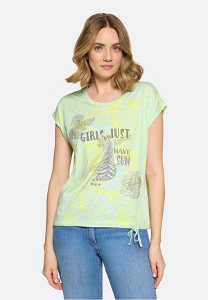 Femme aux cheveux blonds mi-longs portant un t-shirt graphique vert clair avec un zèbre et le texte "Les filles veulent juste du soleil" et un jean bleu.
