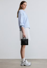 Jeune femme portant un sweat à capuche bleu clair, une jupe plissée blanche, des baskets blanches, tenant un petit sac à main noir, debout devant un fond clair uni.