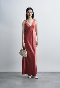 Γυναίκα με αμάνικο κοραλλί μάξι φόρεμα με βαθύ V λαιμόκοψη, κρατώντας μια μπεζ τσάντα χειρός και φοράει λευκά μυτερά τακούνια μπροστά σε έναν απλό τοίχο.