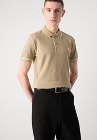 Beige polo krekls ar baltu apdari, ar vienas pogas aizdari, īsām piedurknēm un CK logotipu uz kreisā krūšu, savienots ar melnām biksēm un jostu.