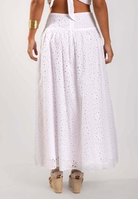 Falda maxi blanca bordada con un diseño acampanado, que presenta patrones florales recortados y una cintura alta. Combinada con sandalias de cuña beige.