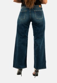 Jeans a gamba larga di un blu scuro con due tasche posteriori, lievi sbiadimenti e dettagli di cucitura. Indossati con scarpe nere con tacco.