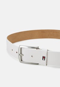 White leather belt with a silver metal buckle, featuring a rectangular logo accent in red, white, and blue. Smooth texture, classic design.