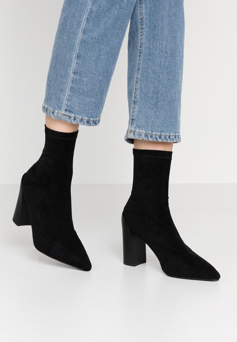 Pieds chaussés de bottines noires en daim à bout pointu avec talons carrés, portées avec un jean bleu clair raccourci, sur un fond uni.