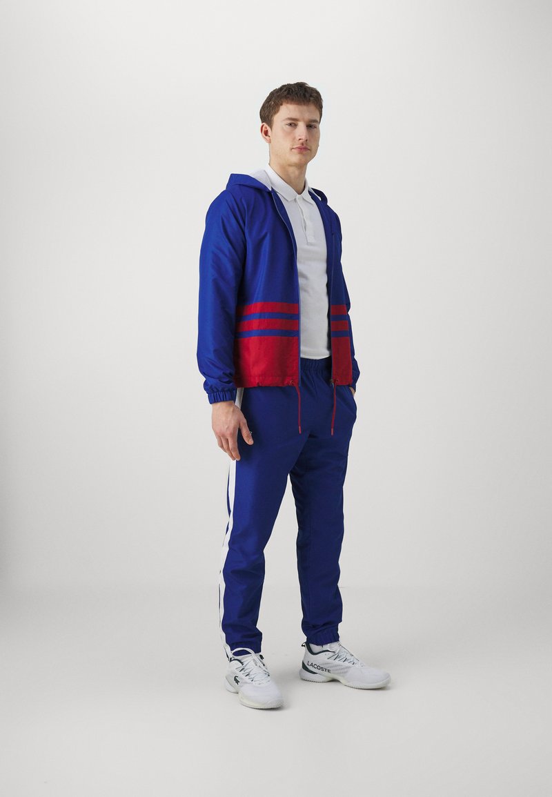 Un giovane che indossa una giacca zip blu e rossa, pantaloni sportivi blu con strisce bianche, una polo bianca e sneaker bianche Lacoste in piedi.