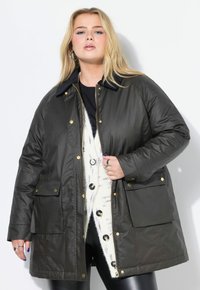 Un lungo cappotto verde oliva scuro con chiusura a zip, accessori dorati e due tasche frontali. Presenta un colletto nero ed è indossato sopra un maglione beige.