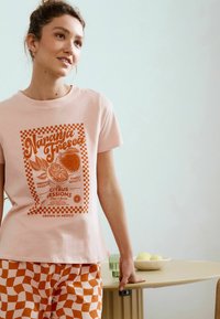 Helles rosa Baumwoll-T-Shirt mit orangefarbener Grafik, die eine halbierte Orange zeigt. Kombiniert mit orange-weiß karierten Hosen.