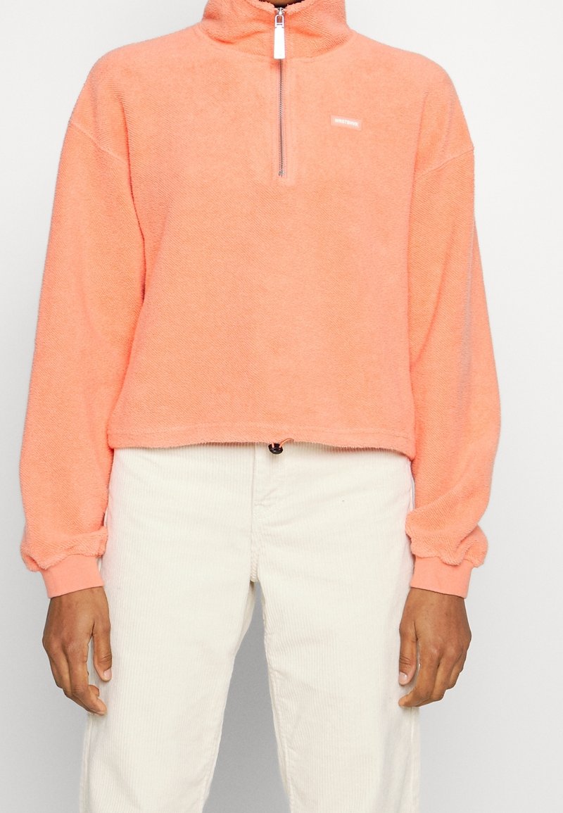 Personne portant un pull en polaire corail à demi-fermeture éclair avec des poignets élastiques et un pantalon écru sur un fond uni.