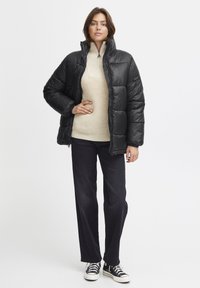 Svart dunjakke med quiltet design, høy krage og glidelåslukking. Bært over en lys beige strikkegenser, kombinert med mørke jeans og joggesko.