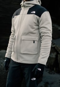 Hellgrau zip-up Jacke mit schwarzen Schulterakzenten, zwei Fronttaschen und Logodetail. Kombiniert mit schwarzen Handschuhen und Hose. Weiche Textur.