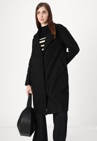 Cappotto nero a trama con rever, maniche lunghe e tasche laterali. Indossato sopra una maglietta a righe bianco e nera. Tiene una borsa nera.