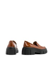 Zapatos de charol marrón tipo mocasín con una gruesa suela de plataforma negra y costuras blancas a lo largo del borde. Diseño de puntera redondeada.