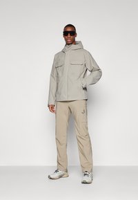 Beige waterdichte jas met twee grote voorzakken, gecombineerd met lichtbeige broek en grijze sportschoenen. Het model draagt een paar donkere zonnebrillen.