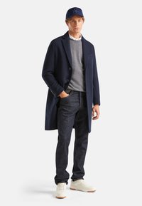 Abrigo de lana navy, suéter gris, jeans de mezclilla azul y zapatillas blancas; presenta una gorra con logo y un corte entallado con detalles de botones.