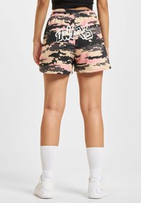 Camouflage-print shorts in pink, black, and beige; elastic waist; bold white text on back; smooth fabric with a relaxed fit.