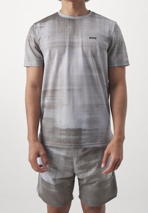 Mężczyzna ubrany w szarą koszulę z krótkim rękawem i wzorem z logo "BOSS" oraz do tego pasujące szorty, stojący na jednolitym tle.