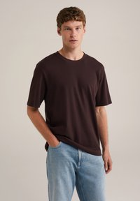 Camiseta de manga corta marrón oscuro en corte relajado, hecha de una tela suave con un cuello redondo clásico. Llevada con jeans de color azul claro.
