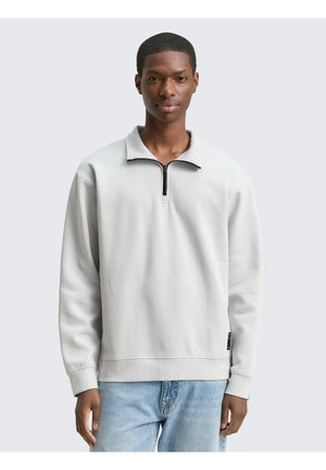 Joven con sudadera gris clara con cremallera hasta la mitad y vaqueros azul claro, de pie frente a un fondo liso gris claro.