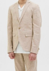 Chaqueta de traje de lino beige claro con cierre de un solo botón, solapas con muesca y dos bolsillos frontales, combinada con una camisa blanca debajo.