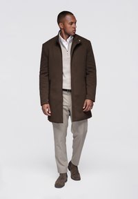 Hnědý dlouhý kabát s vysokým límcem, nošený přes světle svetr a světle zbarvené kalhoty. Má zipy a hladkou texturu. Hnědé kotníkové boty.