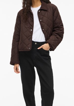 Personne portant une veste matelassée marron foncé sur une chemise blanche, avec un pantalon noir taille haute, une main dans la poche, cadrée au niveau du cou.